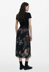 Zwarte ribbeltop gecombineerd met een lange, kleurrijk bedrukte rok met bloem- en paisleypatronen. Het model draagt zwarte sokken en schoenen.