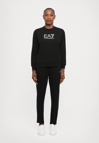 Fekete EA7 Emporio Armani pulóvert és hozzáillő nadrágot viselő személy, egyenes háttérrel, fehér alapon.