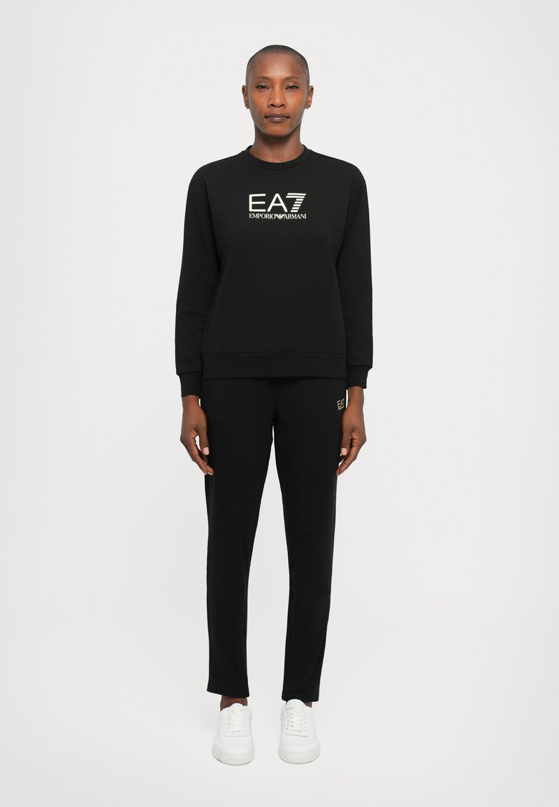 Fekete EA7 Emporio Armani pulóvert és hozzáillő nadrágot viselő személy, egyenes háttérrel, fehér alapon.