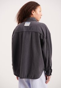 Ženska s kodrastimi lasmi nosi veliko oprano črno denim jakno z belo etiketo na zgornjem hrbtu, združeno s svetlo modrimi hlačami.