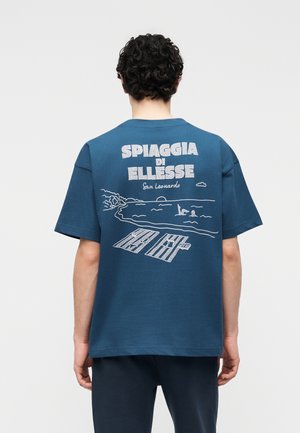 Persoană cu păr creț care poartă un tricou albastru cu un desen alb al unei scene de plajă și textul "SPIAGGIA DI ELLESSE San Leonardo" pe spate.