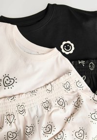 Sort sweatshirt med smiley blomstlogo, lys rosa top med hjerte mønster, og sorte shorts med smilende hjerter, alle lavet af blødt stof.