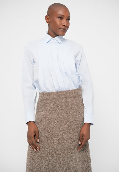 Svetlo modra srajca s pokončnim ovratnikom in nabranim prednjim delom, v kombinaciji z teksturirano rjavo krilo, ki ima visok pas in srednjo dolžino.