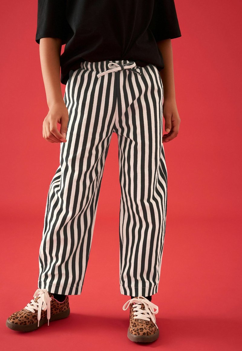 Persona che indossa pantaloni a righe verticali bianche e nere con coulisse e sneakers con stampa leopardata e lacci bianchi su sfondo rosso.