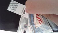 Una mano tiene un'etichetta di abbigliamento con la dicitura "100% cotone biologico" e un'etichetta del marchio Gucci su jeans in denim azzurro.