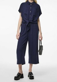 Tuta blu navy con maniche corte, apertura frontale con bottoni, dettaglio in vita con cintura, due tasche sul petto e gambali larghi; abbinata a zoccoli neri con plateau.