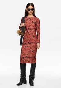 Vestido de manga larga en un patrón salpicado de rojo y negro, hecho de tela suave, combinado con botas altas negras y un bolso pequeño con detalles de piel.