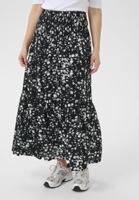 Jupe maxi noire à fleurs avec une taille smockée, ornée de motifs floraux blancs et d'un design en volants, associée à des baskets de couleur claire.