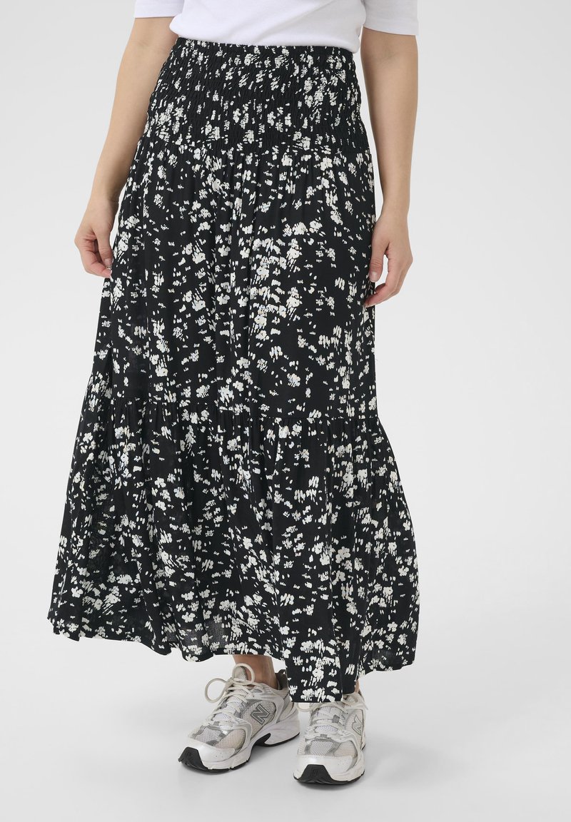 Jupe maxi noire à fleurs avec une taille smockée, ornée de motifs floraux blancs et d'un design en volants, associée à des baskets de couleur claire.