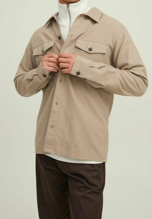 Homme boutonnant une veste-chemise beige sur un col roulé blanc, portant un pantalon marron foncé, sur un fond beige uni.
