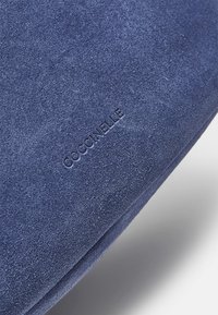 Mörkblå mockamaterial med en slät textur, med präglat varumärkesnamn "COCCINELLE." Designen är platt och minimalistisk.