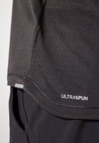Svart ytelsesstoff med teksturert vev, med en etikett merket "ULTRASPU" i hvitt, som viser en sømdetalj på nederkanten.