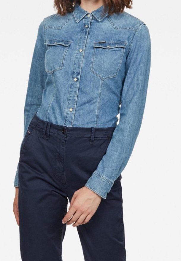 Ljusblå denimskjorta med spetsig krage, långa ärmar, knäppning framtill och två bröstfickor. Kombinerad med marinblå byxor.