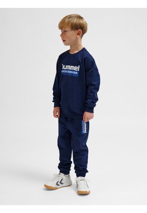 Marineblauwe sweatshirt en joggers met een sportief ontwerp, voorzien van witte en blauwe logo-accenten. De broek heeft elastische boorden en zakken. Witte schoenen met grijze en zwarte details.