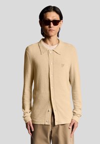 Beige strukturerad cardigan med knappframsidan, spetsig krage och liten broderad logotyp. Bärs över en ljus skjorta, tillsammans med solglasögon.