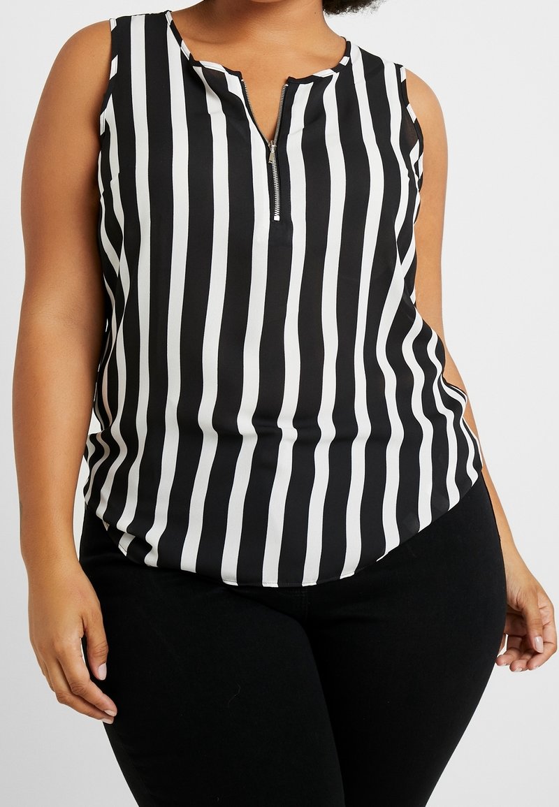 Femme portant un haut sans manches à rayures verticales noires et blanches avec une fermeture éclair à l'avant et un pantalon noir, debout devant un fond uni.
