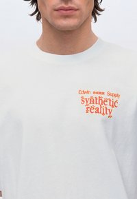 Homme portant un T-shirt blanc avec un texte rouge indiquant "Edwin Synthetic Reality Supply" sur la partie supérieure de la poitrine.