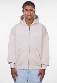 Beżowa bluza z kapturem na zip z dużą kieszenią z przodu, ściągaczami na rękawach oraz regulowanym kapturem. Charakteryzuje się dyskretnym haftem logo na klatce piersiowej.