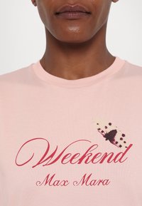 Gros plan d'une personne portant une chemise rose clair avec un texte rouge "Weekend Max Mara" et un petit graphique de papillon.
