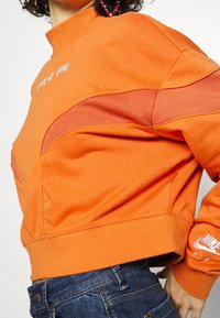 Orange sweatshirt med hög krage, med en kontrasterande paneldesign. Visar "AIR" och Nike-logotypen i vitt på ärmen. Mjuk textur.