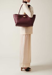 Burgundy leather tote bag with textured finish, gold hardware, and long handles, displayed alongside a light-colored outfit and black heeled shoes.