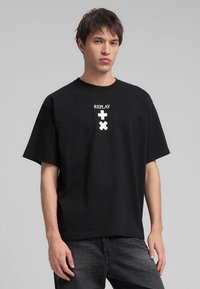 Czarny bawełniany t-shirt z krótkim rękawem, z białym napisem "REPLAY" i graficznymi elementami na klatce piersiowej. Luźny krój, prosty design.