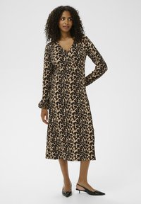 Robe en imprimé léopard beige avec des taches noires. Manches longues, décolleté en V et longueur mi-mollet. Portée avec des escarpins noirs à bout pointu.