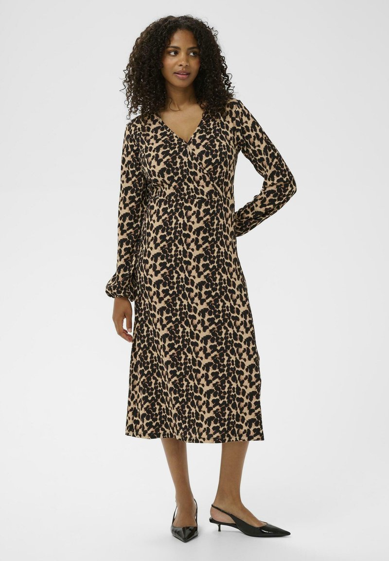 Robe en imprimé léopard beige avec des taches noires. Manches longues, décolleté en V et longueur mi-mollet. Portée avec des escarpins noirs à bout pointu.