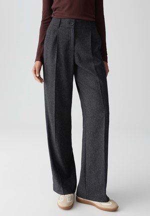 Vrouw in een donkergrijze wijde broek, bruin ribbeltrui met lange mouwen en beige sneakers, staande tegen een effen achtergrond.