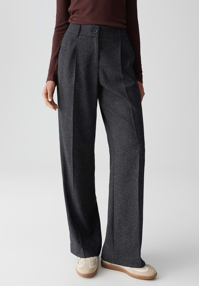 Femme portant un pantalon large gris foncé, un haut à manches longues côtelé marron et des baskets beiges, se tenant contre un fond uni.