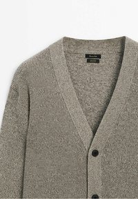 Grå stickad cardigan med V-ringning. Har ett strukturtyg med ett fint mönster och svarta knappar framtill.