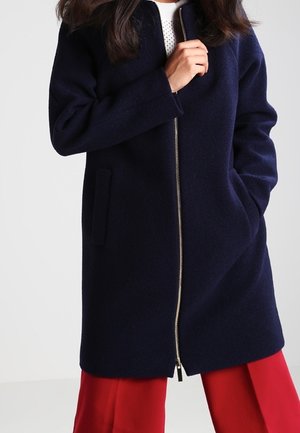 Kobieta ubrana w granatowy płaszcz na zamek z bocznymi kieszeniami, na białej bluzce i jasnoczerwonych, szerokich spodniach, pozuje z jedną ręką w kieszeni.