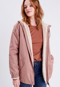 Ljusrosa parka med foder av konstpäls, dragkedja, sidofickor och elastiska muddar, bärs över en strukturerad brun topp.