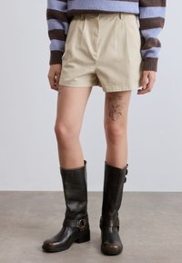 Μπεζ σορτς σε στενή γραμμή με πιέτες, φτιαγμένα από ύφασμα με μείγμα βαμβακιού. Συνδυάζονται με καφέ δερμάτινες μπότες μέχρι το γόνατο με λουράκια και αγκράφες.