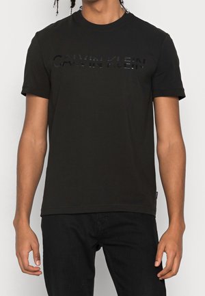 Person trägt ein schwarzes Calvin Klein T-Shirt mit kurzen Ärmeln und dezenter glänzender Logo-Prägung auf der Brust, kombiniert mit schwarzen Hosen.
