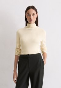 Pull en maille côtelée, crème, à col roulé et manches longues, associé à un pantalon noir taille haute, affichant un design simple et ajusté.