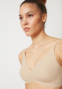 Beige bra with a smooth texture, V-neck design, and wide straps. Subtle branding near the bottom right corner.