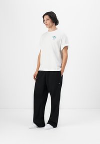 Hvit bomull T-skjorte med rullede ermer og et lite blått grafisk design, kombinert med løstsittende svarte bukser og hvite joggesko.