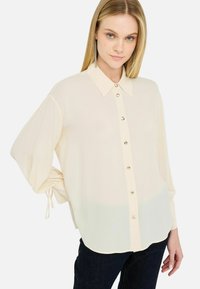 Helle Cream-Bluse mit einem strukturierten Kragen, vorderen Knöpfen und gerafften Ärmeln; aus weichem, transparentem Stoff mit glatter Textur gefertigt.