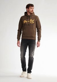 Felpa marrone con scritta gialla, coulisse e tasca frontale, abbinata a jeans scuri e scarpe da ginnastica bianche, in piedi contro uno sfondo chiaro.