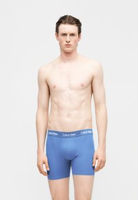 Νέος άνδρας με ανοιχτόχρωμο δέρμα και καστανά μαλλιά στέκεται με πρόσωπο προς τα εμπρός, φοράει μπλε μπόξερ της Calvin Klein σε λευκό, απλό φόντο.