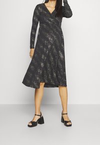 Svart omlottklänning med långa ärmar och ett mönster av metalliskt glittter. Utställd kjol som når till mitt på vaden, tillsammans med svarta klacksandaler.