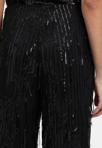 Zwarte broek met een gelaagd ontwerp en cascaderende pailletten die een gestructureerd, glinsterend effect creëren. Close-up toont duidelijke franjes. 