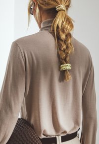 Langärmliges, taupefarbenes Rollkragenoberteil mit weicher Textur. Haar in einem Zopf gestylt, mit einer goldenen Haarklammer fixiert. Braune geflochtene Tasche sichtbar.