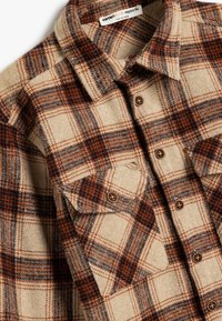Camisa de franela con un patrón de cuadros en beige, naranja y azul marino; cuenta con dos bolsillos en el pecho y cierres de botón marrón. Tejido suave y texturizado.
