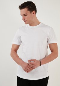 Fehér pamut póló rövid ujjal, kerek nyakkal és diszkrét szegéllyel. Fekete nadrággal viselve, kezek elöl helyezve.
