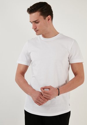 T-shirt alb din bumbac cu mâneci scurte, guler rotund și detalii subtile la tiv. Purtați cu pantaloni negri, mâinile poziționate în față.
