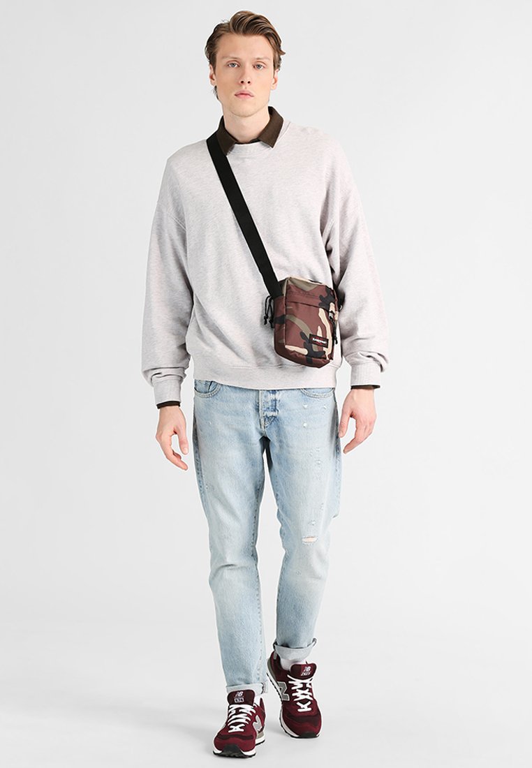 Felpa grigia chiara con colletto marrone, abbinata a jeans chiari sfilacciati. Borsa a tracolla camouflage marrone e sneaker scure completano l'outfit.