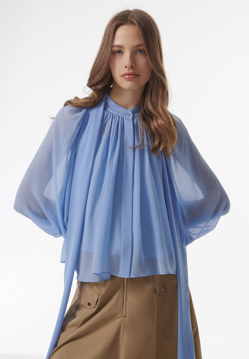Blouse légère bleue claire à encolure haute, avec un devant plissé et des manches fluides. Portée sur une jupe beige avec des zips et des poches.