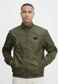 Olive green zippered jacket with a stand-up collar, elastic cuffs, side pockets, and a small black logo patch on the chest. Smooth, lightweight fabric.